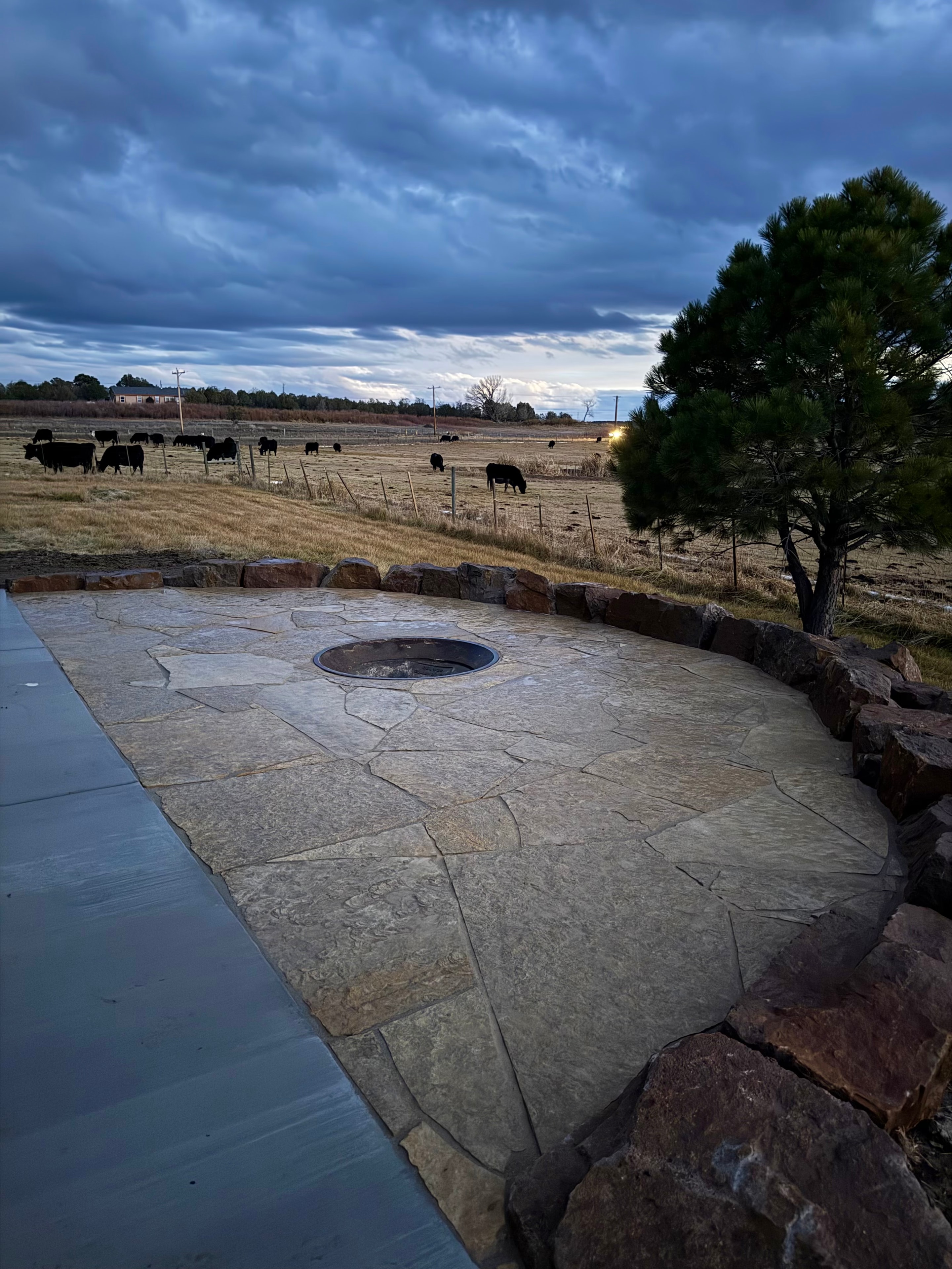 Natural tan flagstone patio with moss rock border and recessed metal fire pit under dramatic stormy sky in Farmington NM — High Desert NativeScapes